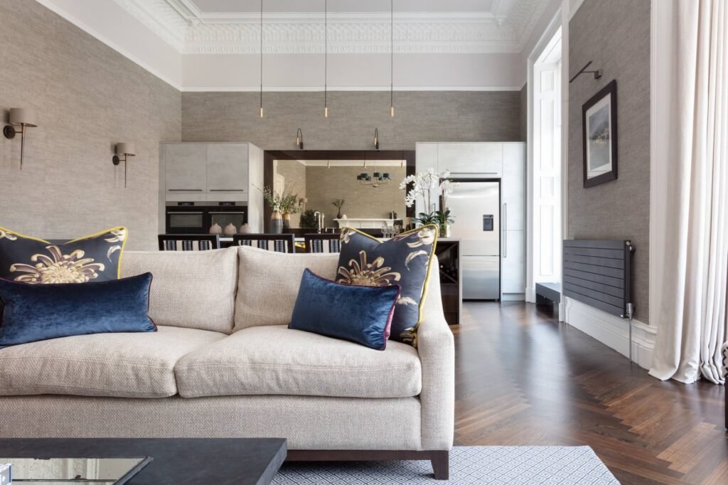 Elegant open-plan living room and kitchen with beige sofa, blue cushions, grey textured walls, and dark herringbone floor.