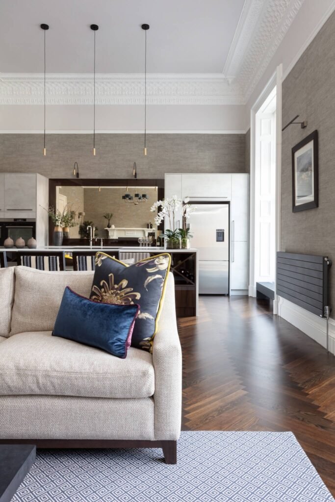 Stylish open-plan living room with beige sofa, blue rug, dark herringbone floor, grey walls, white kitchen cabinets & modern lighting.