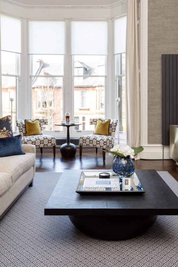 Bright, modern living room with a bay window, patterned armchairs, cream sofa, geometric rug, and decorative black coffee table.