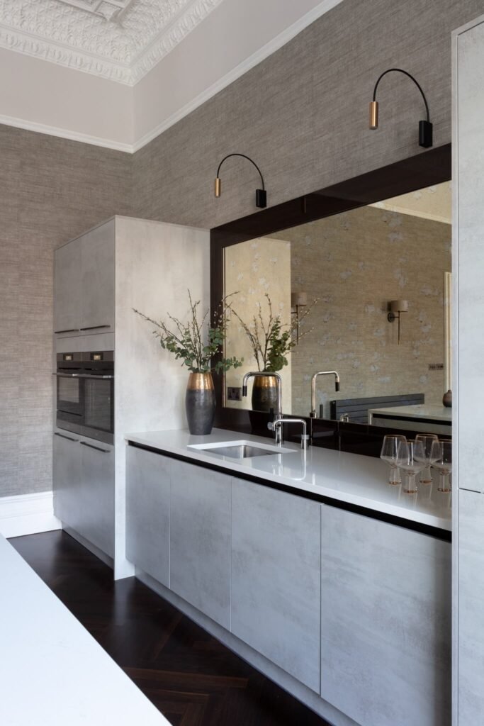 Contemporary kitchen with textured grey cabinets, white countertop, dark herringbone floor, ornate ceiling, and reflective mirror.