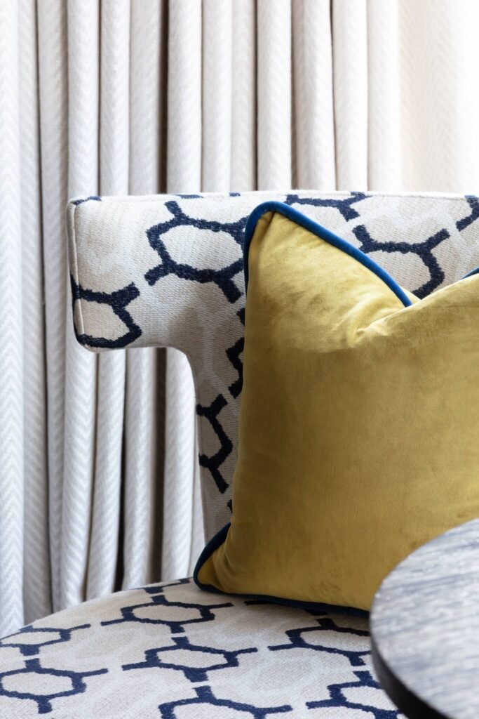 Cream armchair with navy geometric pattern, a yellow velvet pillow with blue piping, and neutral herringbone curtains.