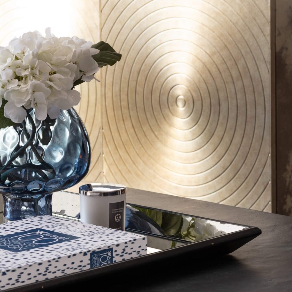 Blue textured glass vase with white hydrangeas, mirrored tray with candle and book, against an elegant circular wall panel.