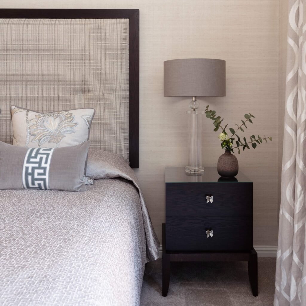 Elegant neutral bedroom with framed upholstered headboard, decorative pillows, dark wood nightstand, clear lamp, and textured walls.