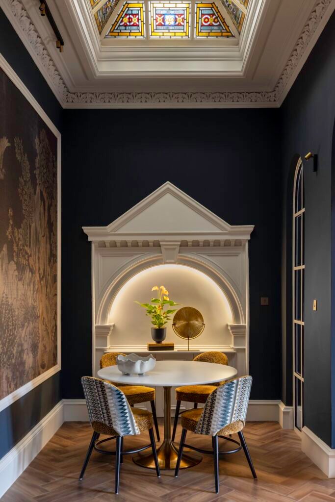 Ornate dark blue dining nook with white arched alcove, round table, modern chairs, stained glass skylight, herringbone floor.