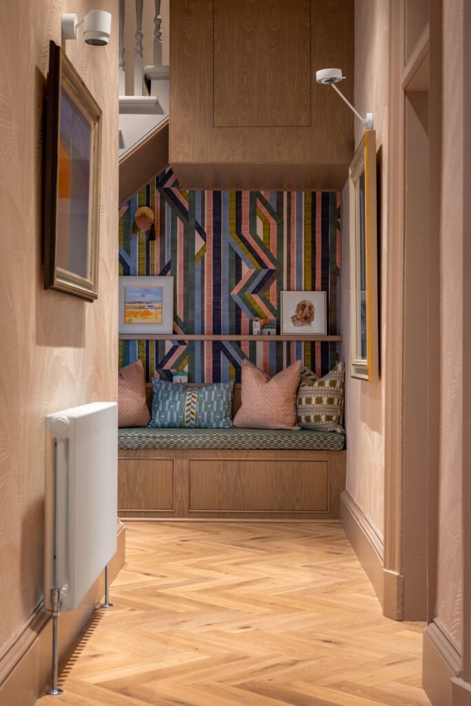 Eclectic hallway with built-in reading nook under stairs, colorful geometric wallpaper, patterned pillows, and herringbone wood floor.