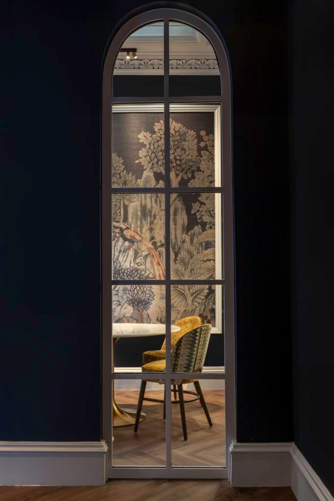 Arched frame revealing a dark room with a framed nature mural, round table, patterned chairs, and ornate ceiling.