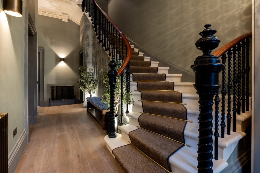 Grand traditional foyer with sweeping staircase, herringbone runner, ornate dark balusters, and warm lighting.