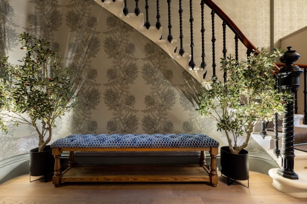 Stylish hallway with thistle wallpaper, blue tufted bench, two olive trees, and a dark ornate staircase.