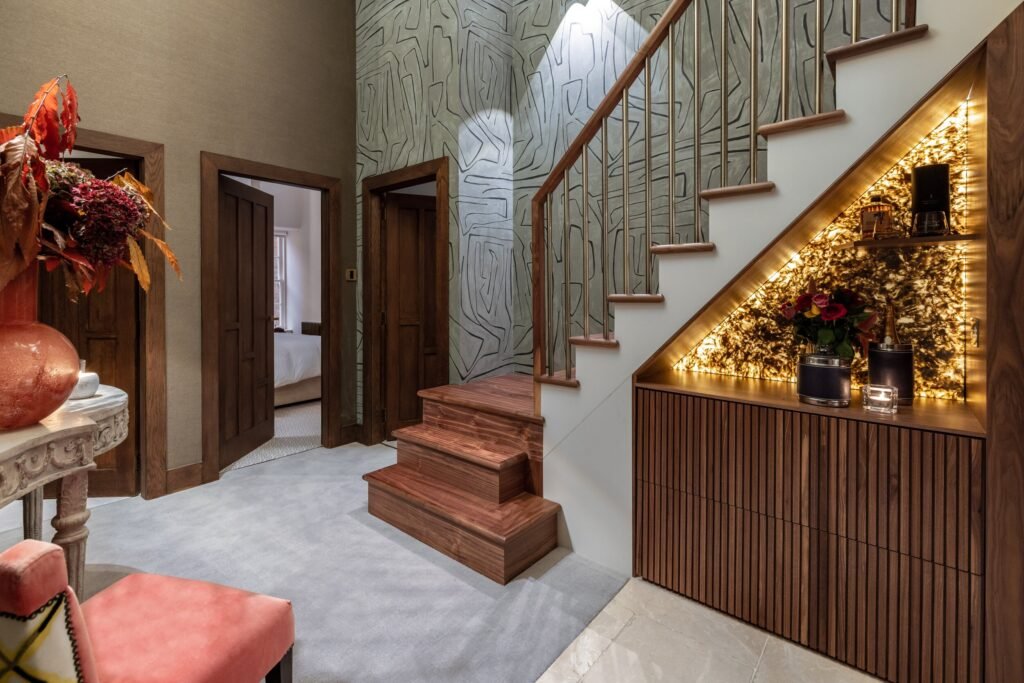Luxury entryway with wooden staircase, brass balusters, abstract patterned wallpaper, and an illuminated stone under-stair display.