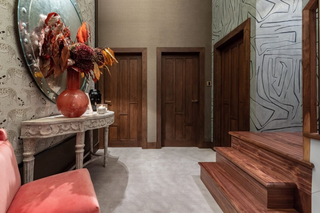 Eclectic entryway with ornate console, mirror, fall decor, leopard and abstract wallpapers, dark wood doors, and wood stairs.