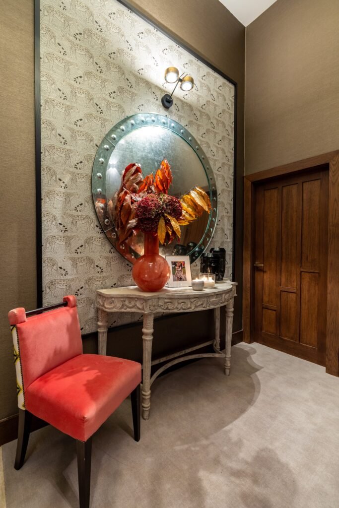 Stylish entryway with leopard wallpaper, round studded mirror, ornate console table with fall decor, and a vibrant coral chair.