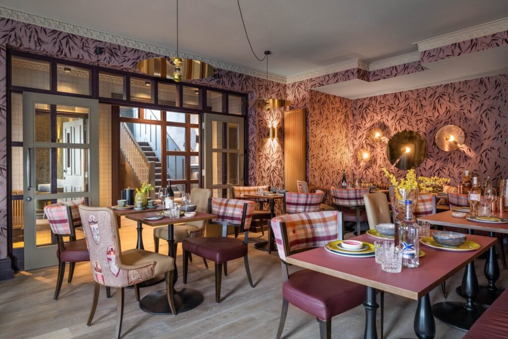 Chic restaurant interior featuring purple bamboo wallpaper, plaid and leather chairs, gold accents, and set tables.