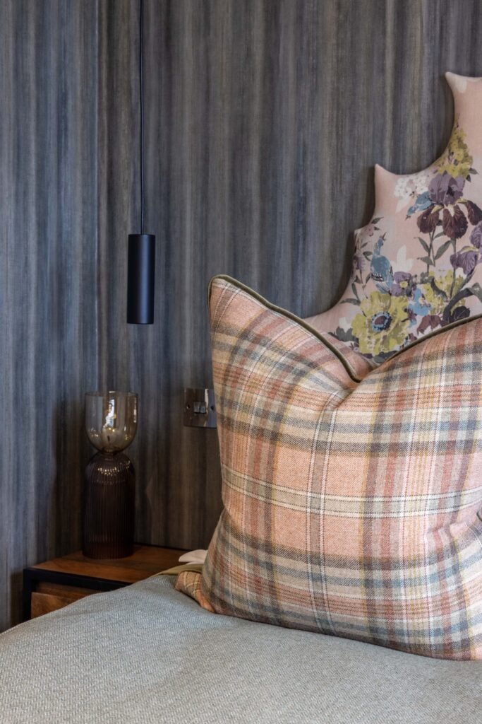Modern bedroom detail: Warm plaid pillow, floral headboard, textured grey wall, and sleek black pendant light.