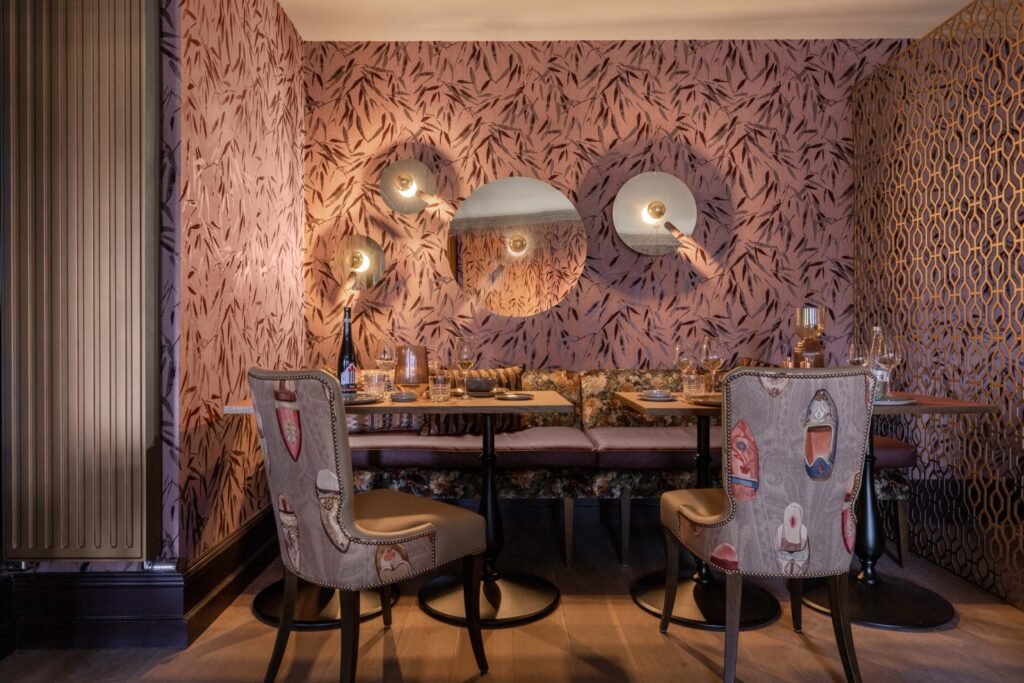 Intimate dining room with blush bamboo wallpaper, eclectic chairs, plush booth seating, modern wall sconces, and a gold geometric divider.