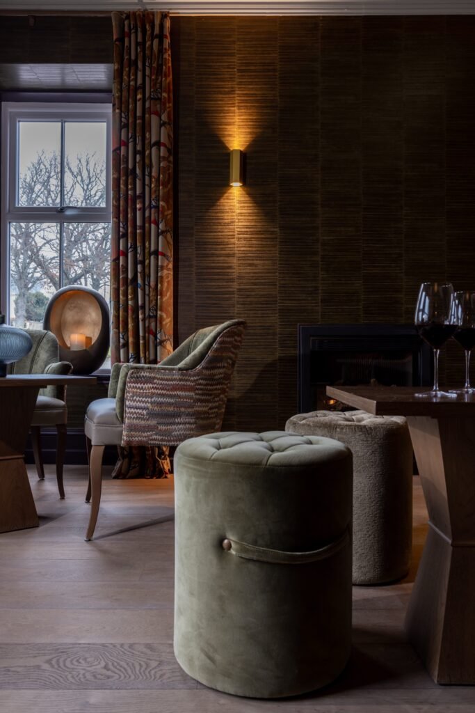Cozy, elegant lounge with dark textured walls, patterned curtains, velvet armchairs and ottomans, warm lighting, and a fireplace.