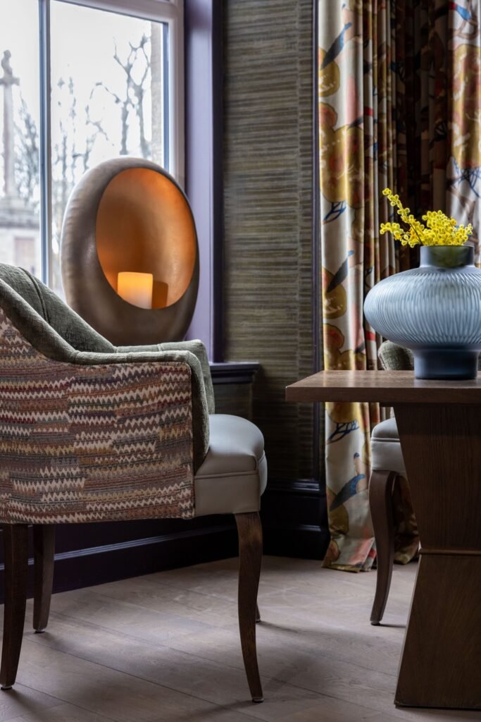 Elegant interior with patterned armchair, candle in golden holder, floral curtains, textured wall, and mimosa in blue vase.