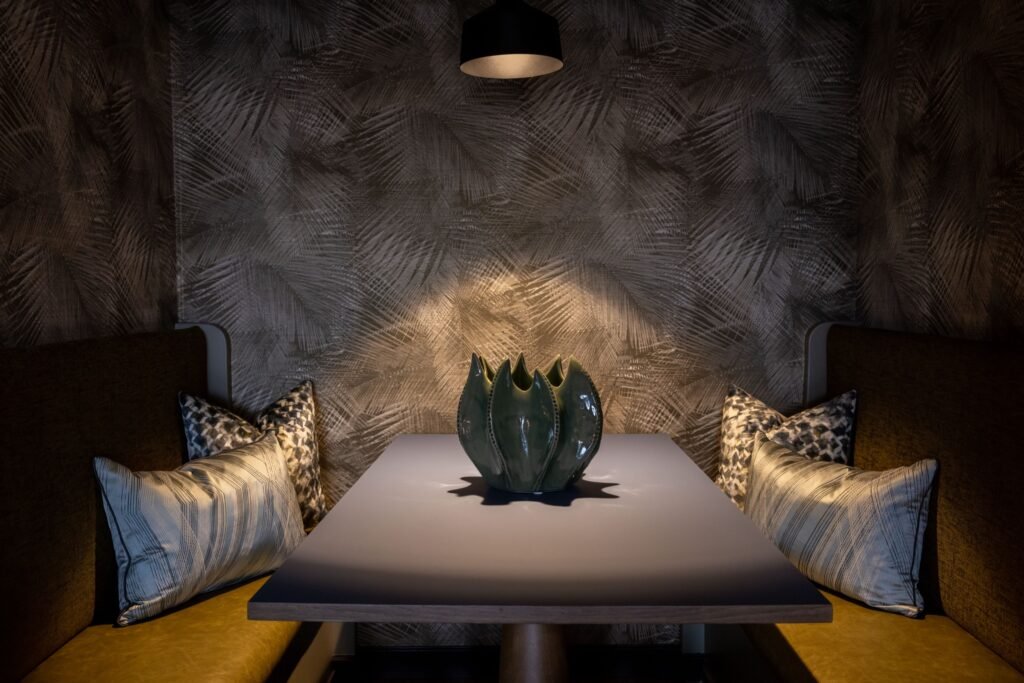 Cozy dining booth with dark palm leaf wallpaper, pendant light, and a striking green vase on a modern table.