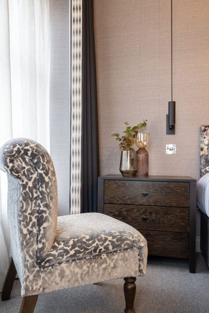 Stylish bedroom corner: grey leopard print chair, dark nightstand, modern wall light, textured wallpaper, patterned curtains.
