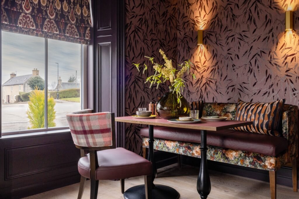 Inviting dining nook with dark leaf wallpaper, floral banquette, plaid chair, pink table, and warm wall lights.