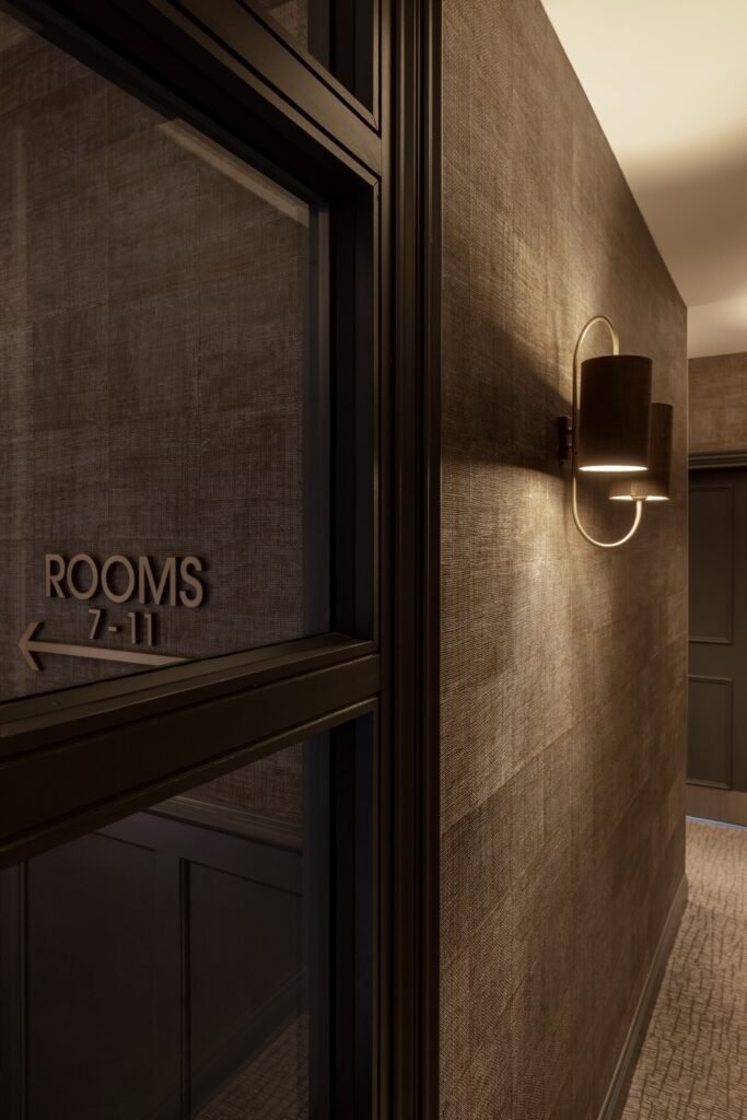 Modern hotel hallway design with bronze textured walls, glowing sconce, and 'Rooms 7-11' sign on frosted glass.
