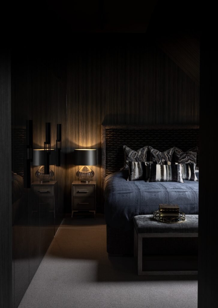 Dark luxury bedroom with blue quilted bedding, patterned pillows, and a warm bedside lamp.