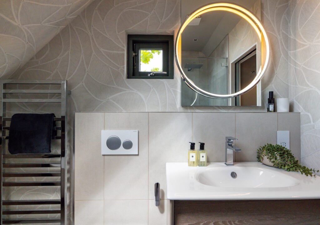 Bright modern bathroom with organic patterned wallpaper, lit round mirror, white vanity, & chrome towel warmer.