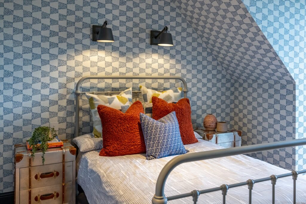 Cozy attic bedroom with blue geometric wallpaper, grey metal bed, rust and patterned pillows, vintage trunk nightstands, and wall sconces.