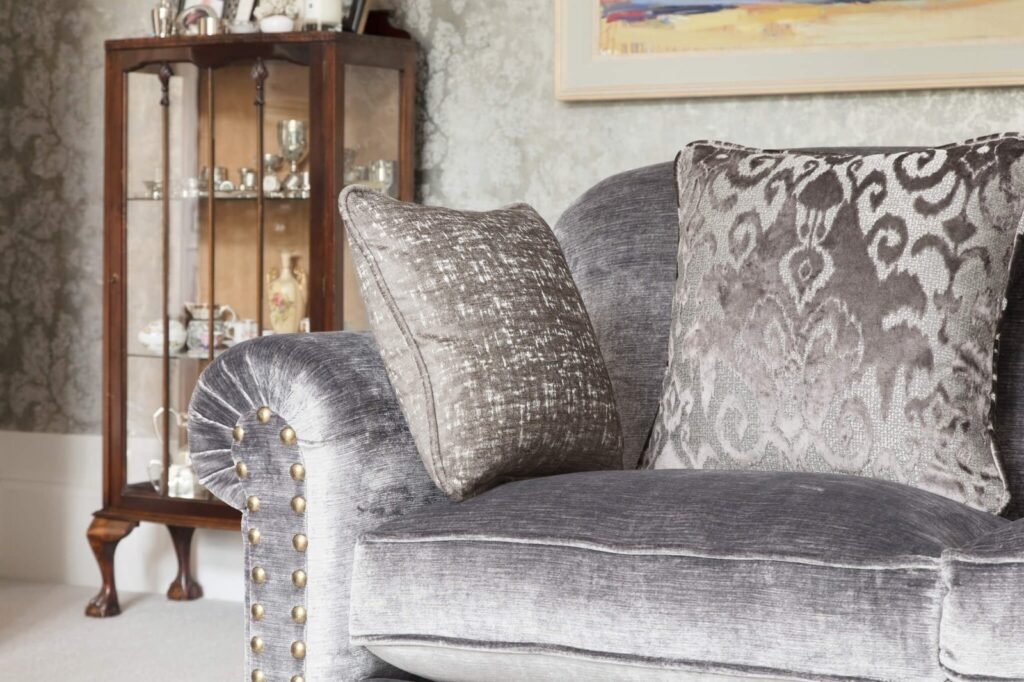 Luxurious grey velvet sofa with brass nailhead trim and ornate patterned cushions, set in a traditional living room.