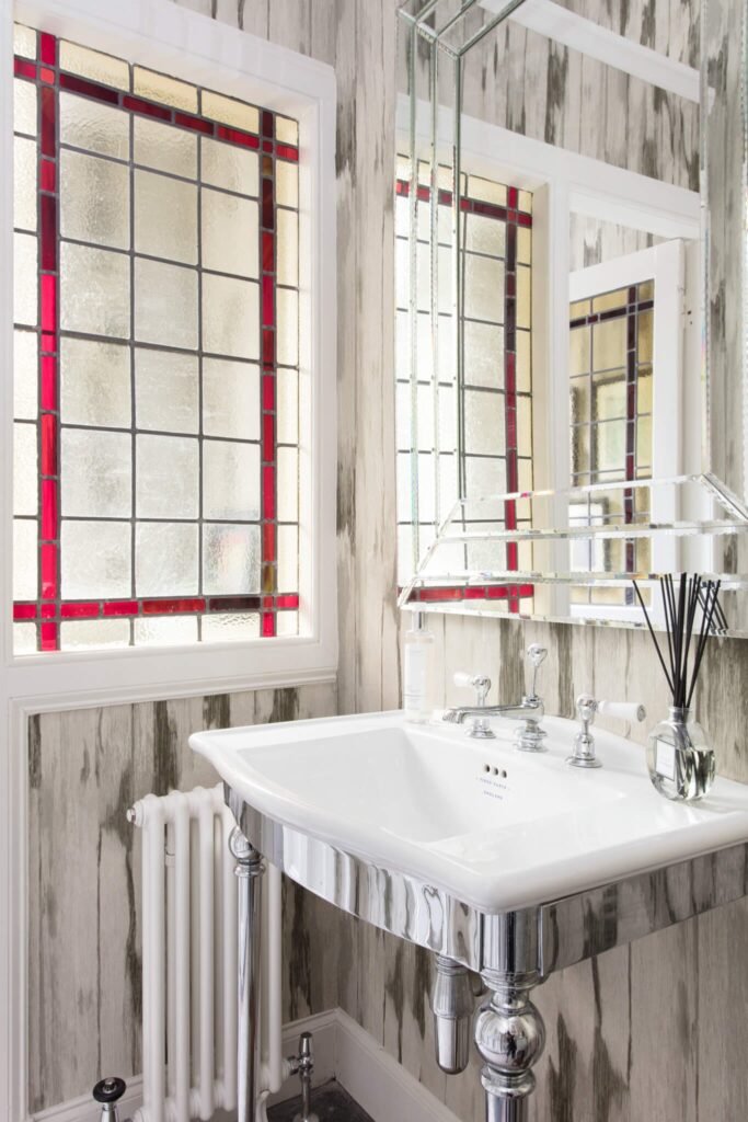 Elegant bathroom with a red stained glass window, white pedestal sink, tiered mirror, and distressed wood-panel wallpaper.