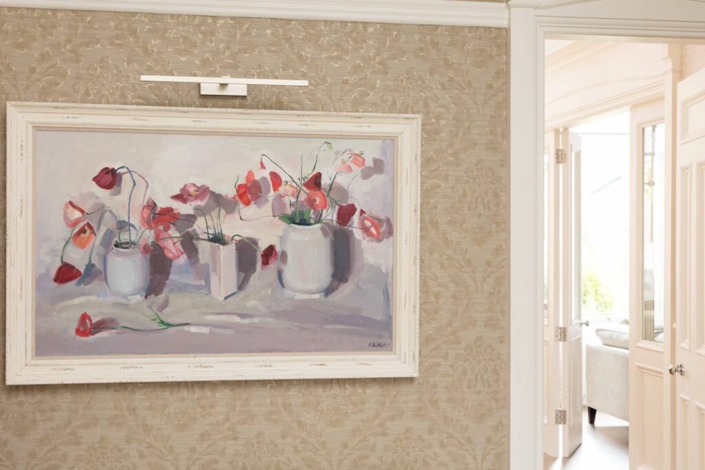 Floral painting with picture light on patterned beige wallpaper. White double doors lead to a bright room.