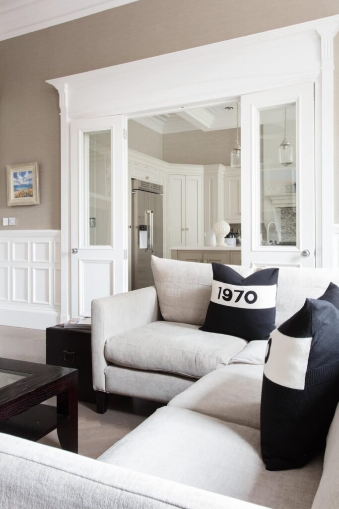 Transitional living room with light sectional, 1970 pillow & wainscoting, opening to a bright white kitchen via French doors.