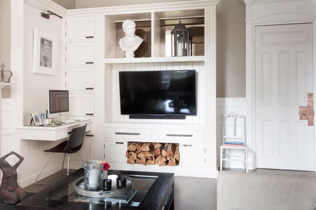 Modern living room with white built-in unit: TV, workstation, display shelves with bust, and firewood storage.