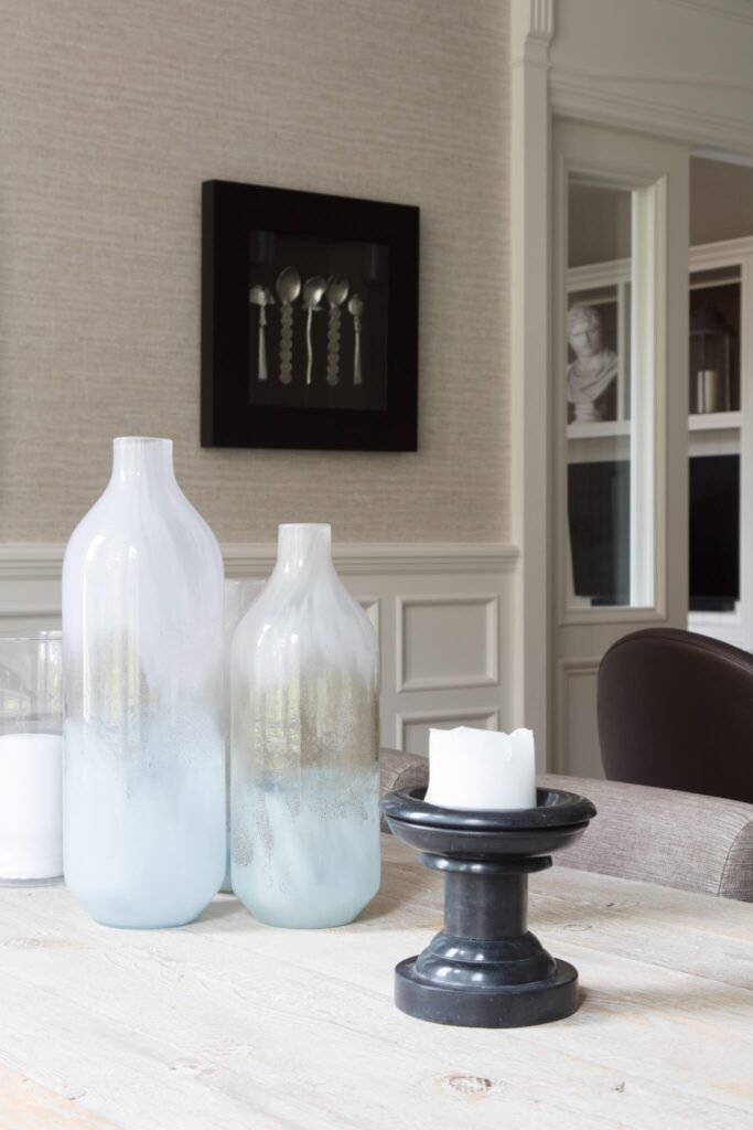Close-up of a light distressed wood table with two frosted blue/white glass vases, a black candle holder, and framed utensil art.