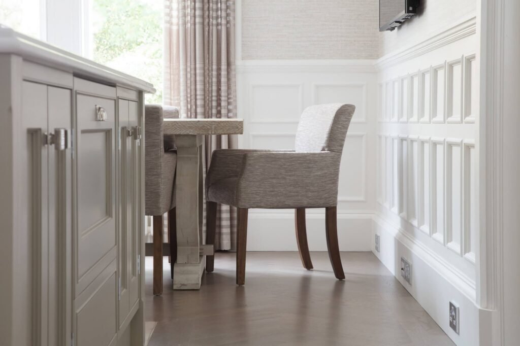 Elegant neutral kitchen dining with grey cabinetry, rustic table, upholstered chairs, white paneled walls & sheer curtains.