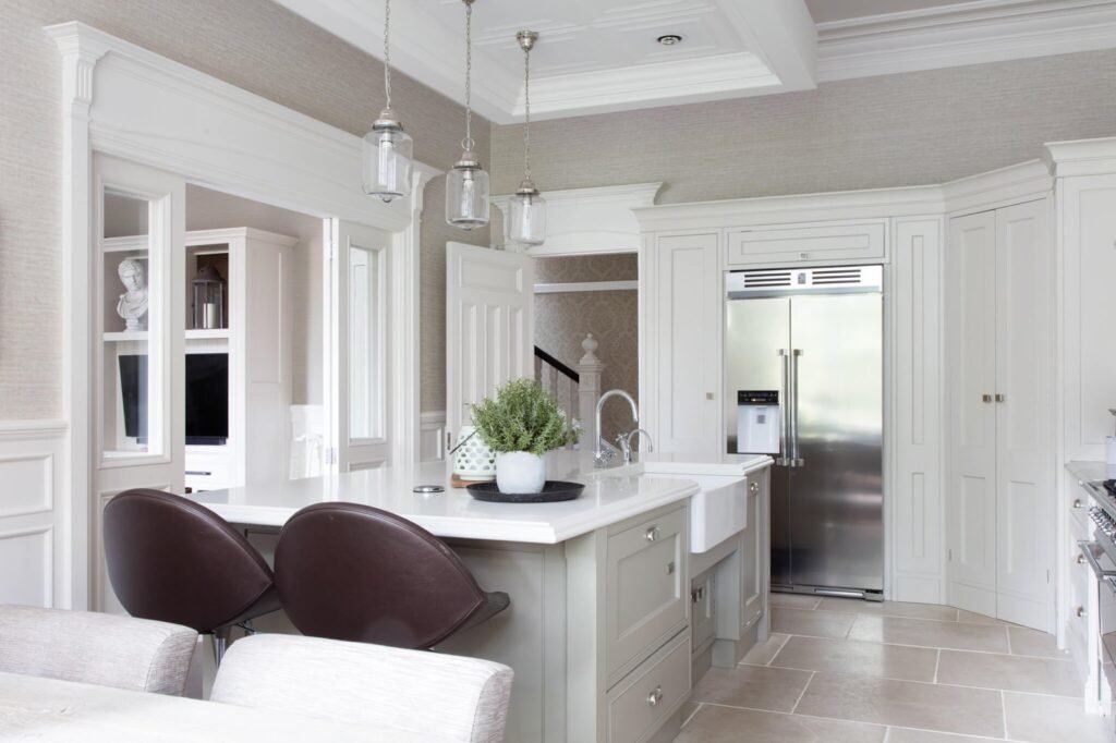 Elegant, traditional kitchen with a greige island, white farmhouse sink, pendant lights, stainless steel fridge, and stone floor.