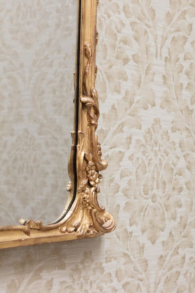 Close-up of a richly ornate gold mirror frame with baroque-style carvings beside beige floral wallpaper.