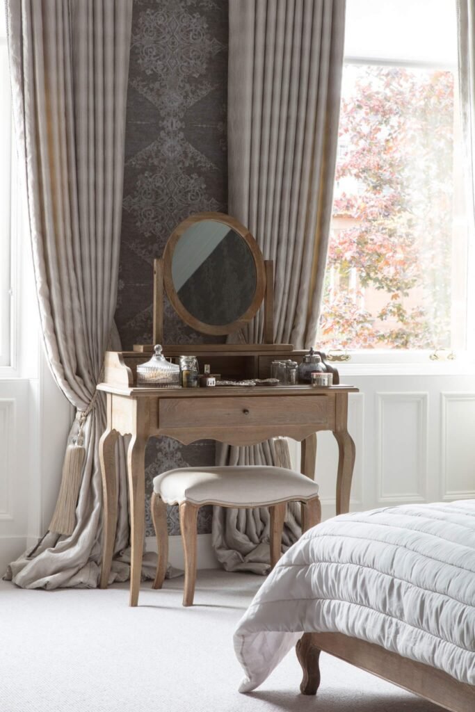 Chic bedroom with light wood dressing table & mirror, stool, neutral curtains, patterned wallpaper, and sunlit autumn window.