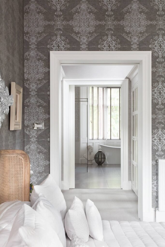 Elegant bedroom with grey damask wallpaper & white bedding, opens to a bathroom featuring a freestanding tub & classical bust.