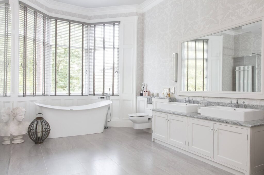 Elegant white bathroom with freestanding tub, bay window with blinds, and double vanity with vessel sinks. Light and spacious.