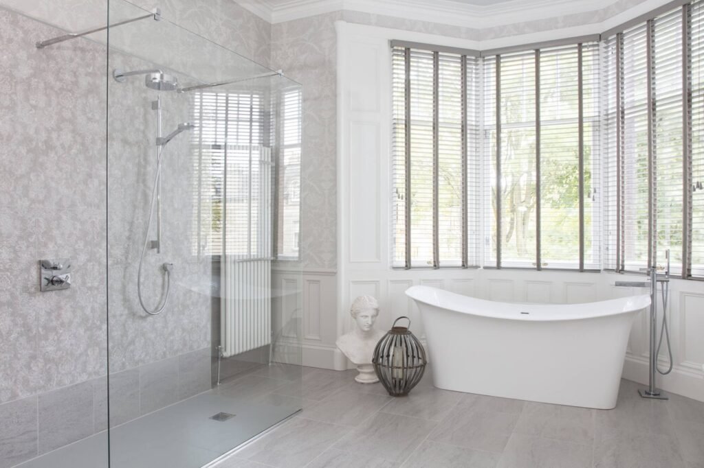 Luxurious grey bathroom with freestanding tub, glass walk-in shower, patterned walls, and bay window blinds.