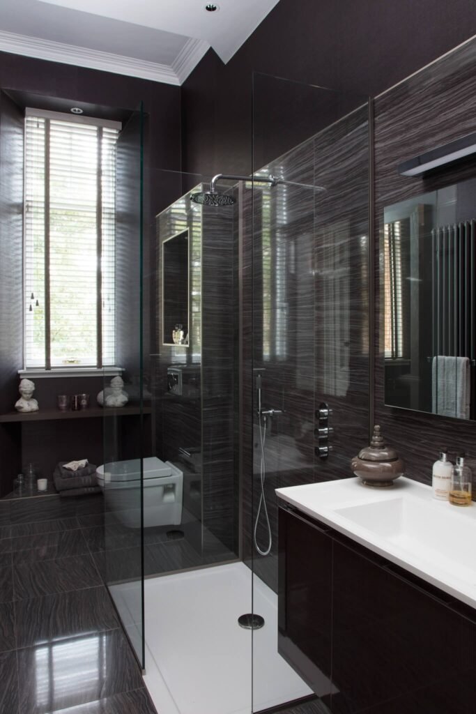Sleek modern bathroom with dark textured tiles, glass walk-in shower, floating vanity, wall-mounted toilet, and window.