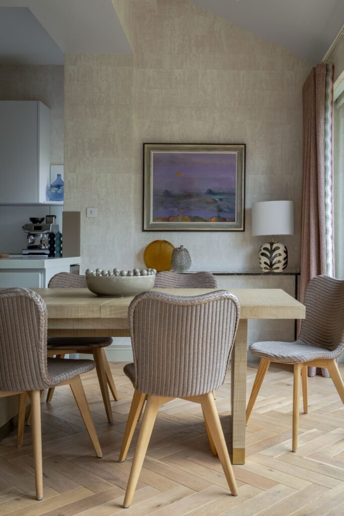 A modern dining area with a light wood table, woven chairs, herringbone floor, neutral textured wallpaper, and abstract art.
