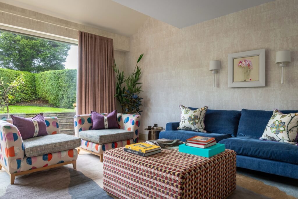 Eclectic living room with blue sofa, patterned armchairs, colorful ottoman, textured walls, and lush garden view.