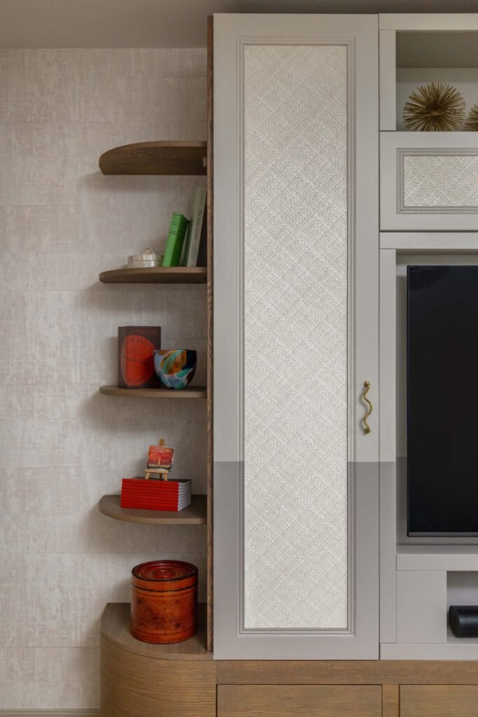 Elegant built-in wall unit featuring curved wooden shelves with decor, light grey cabinetry with woven panels, and an integrated TV.