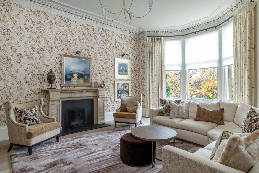 Elegant living room with botanical wallpaper, ornate molding, bay window overlooking fall trees, cream sectional sofa, and fireplace.