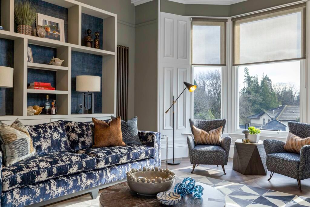 Stylish lounge with blue abstract sofa, built-in shelves, two armchairs, patterned rug, and bright bay window view.