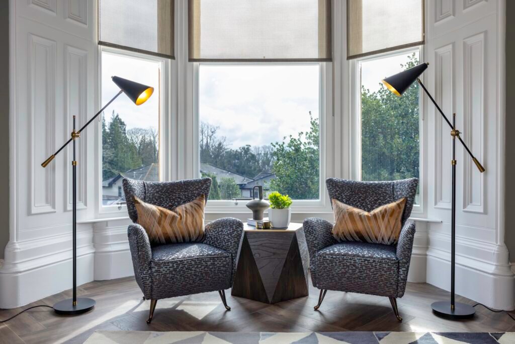 Stylish bay window seating with two patterned armchairs, geometric side table, modern floor lamps, and serene outdoor view.