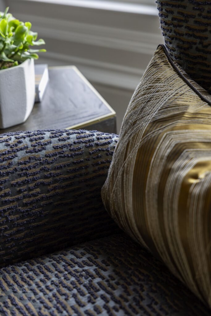 Intricate detail of modern interior textiles: blue textured chair fabric, gold geometric pillow, with plant on side table.