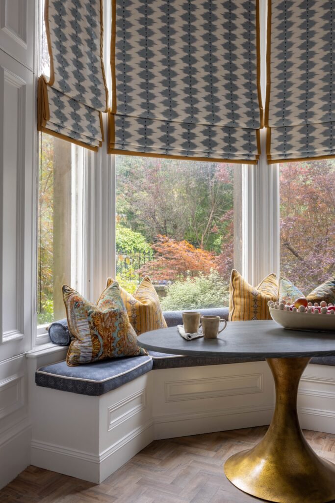 Cozy bay window banquette with ikat Roman blinds, colorful pillows, a dark round table, and a vibrant garden view.