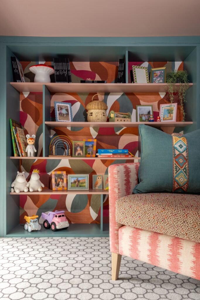 Children's room: Teal built-in shelves with pink accents & abstract wallpaper, full of books, toys, & framed art. Armchair.